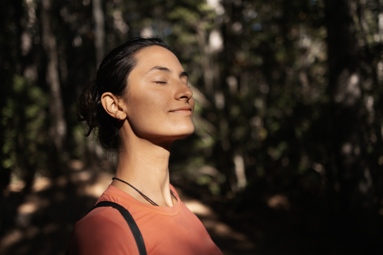 A woman stands outdoors in a forest, enjoying the sunlight with closed eyes and a peaceful expression, conveying tranquility and connection to nature. She is wearing comfortable clothing in a relaxed environment.